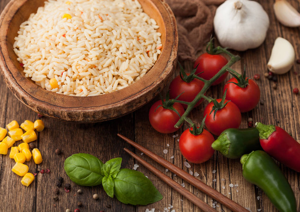 Wooden bowl with boiled long grain basmati rice with vegetables on wooden table background with sticks and tomatoes with corn,garlic and basil. Macro - Photo, Image