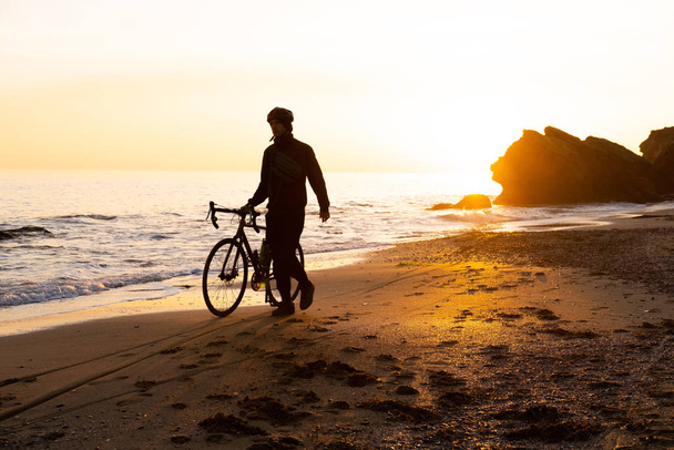 Gün batımında kumsalda kasklı bisiklet süren genç bir erkeğin silueti. - Fotoğraf, Görsel