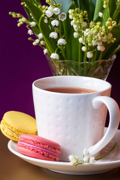 French multicolored cakes macarons and a cup of tea. A bouquet of lily of the valley in a crystal vase. - Photo, Image