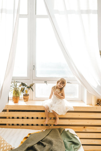 Cute little girl in beautiful dress is sitting on a window sill at light sunny bedroom - 写真・画像
