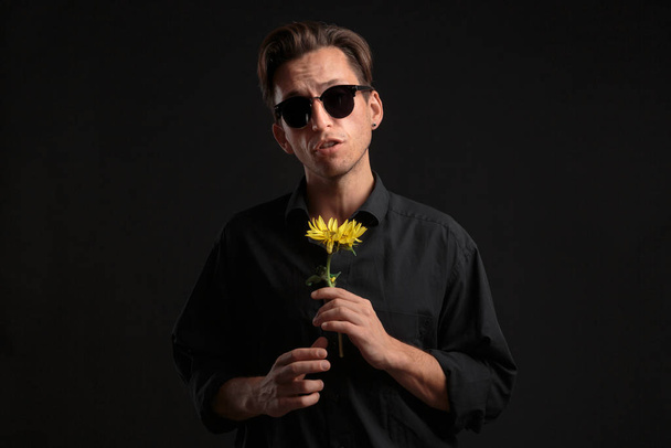 Sexy man in a black shirt and sunglasses holding beautiful yellow flower to give it to his beloved. - Foto, imagen