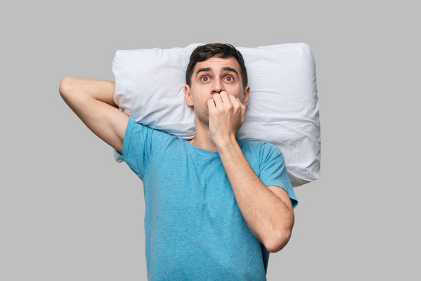 Tired brunet man in a blue tee is scared lying on a white pillow isolated over grey background. - Photo, Image