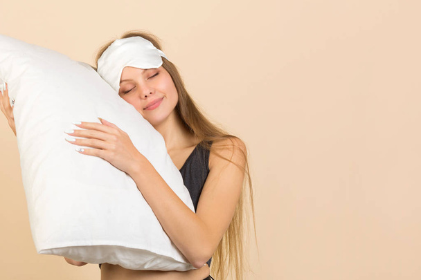 όμορφη νεαρή γυναίκα σε μπεζ φόντο με μάσκα ύπνου με ένα μαξιλάρι στα χέρια της - Φωτογραφία, εικόνα