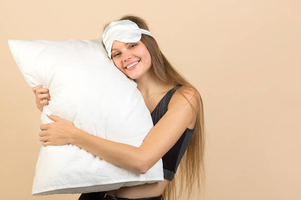 beautiful young woman on a beige background in a sleep mask with a pillow in her hands - Fotó, kép