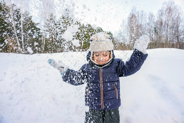 πορτρέτο ενός ευτυχισμένου παιδιού αγόρι ρίχνει χιόνι, νιφάδες χιονιού στον αέρα το κρύο χειμώνα στο φόντο των χιονοστιβάδων - Φωτογραφία, εικόνα