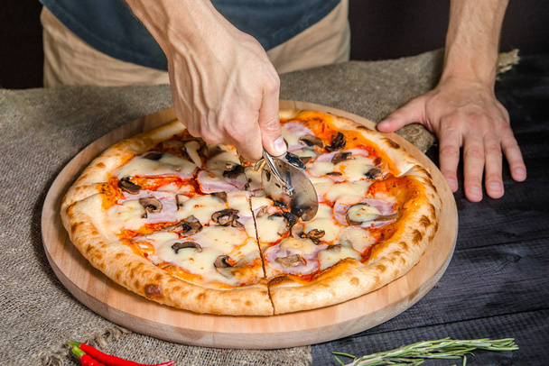 Cortar pizza picante con salami, carne, jamón, salchicha, pimientos, champiñones, tomate, queso con cortador en una tabla de madera
 - Foto, Imagen