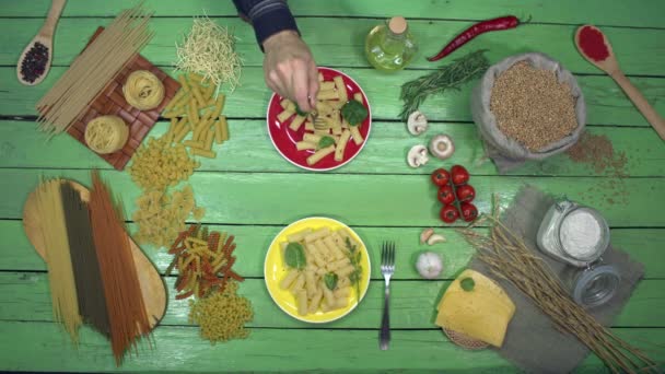 Pasta auf bunten Tellern mit Spinat. Ansicht von oben. Der grüne Tisch ist in viele Arten trockener Pasta und Teig auf der linken Seite und Gemüse, Kirschtomaten, Käse, Aroma und Pilze auf der rechten Seite unterteilt.. - Filmmaterial, Video