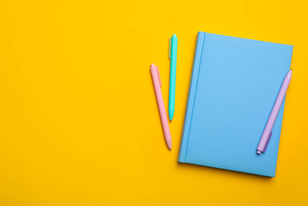 Светло-голубая тетрадь и ручки на желтом фоне, плоские лежат. Пространство для текста
 - Фото, изображение