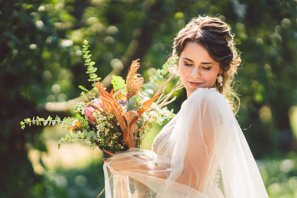 Beautiful bride with bouquet flowers stands on forest background. Rustic style. beautiful bride in delicate dress outdoors. Close up portrait of young bride in park in sunny weather wooded area - Photo, Image