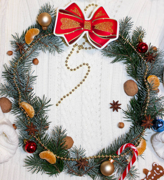 Corona de Navidad de pino decorado con nueces, avellanas, mandarinas, lazo rojo, cuentas de oro, anís estrellas y también bolas, bastón de caramelo y el oso de jengibre en un fondo de punto blanco.Año Nuevo, Flat Lay
 - Foto, imagen