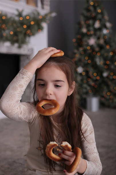 Beautiful girl with long dark hair is fooling around with cookies on New Year's bokeh lights at a decorated Christmas tree - Photo, Image