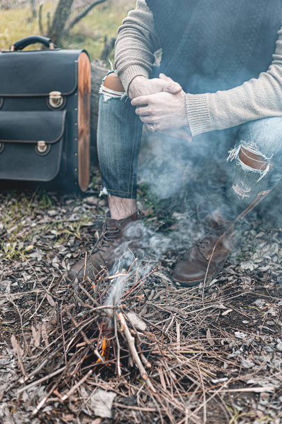 A stock photo of a man in nature sitting by a small fire, warming himself. - Photo, Image