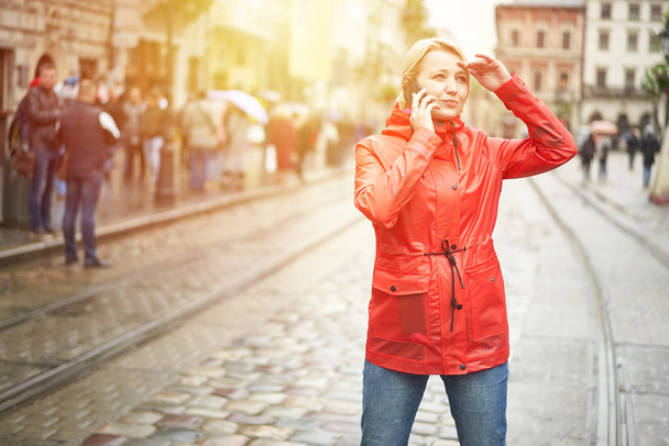 Girl talking on the phone on the street. Portrait of pretty smiling young woman in red bright raincoat. Sunny raining day in city. Image with flare - Photo, image