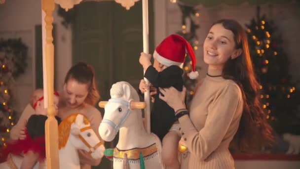 Christmas concept - Two babies sitting on the carousel horses - their smiling mothers standing near by them and holding them - Video, Çekim