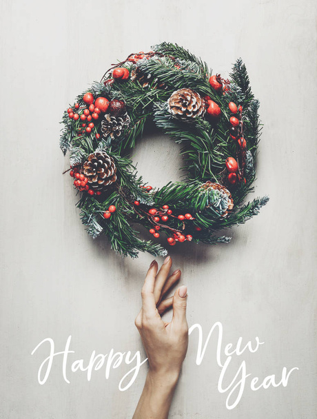 Home decoration for winter holidays. Female hand carefully touches a Christmas wreath on white background, wooden top view. New year concept - Foto, Imagen