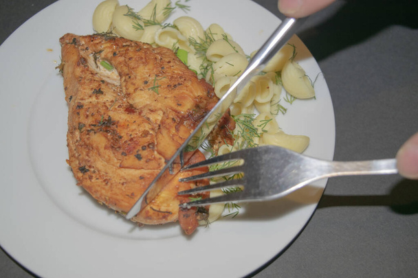 Pechuga de pollo frita con especias, pasta hervida con hierbas, tenedor de metal y cuchillo que cortan un pedazo de la pechuga en un plato blanco
. - Foto, imagen