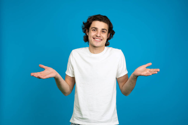 Vue de face du jeune homme souriant montrant un geste d'haussement d'épaules isolé sur fond bleu
 - Photo, image
