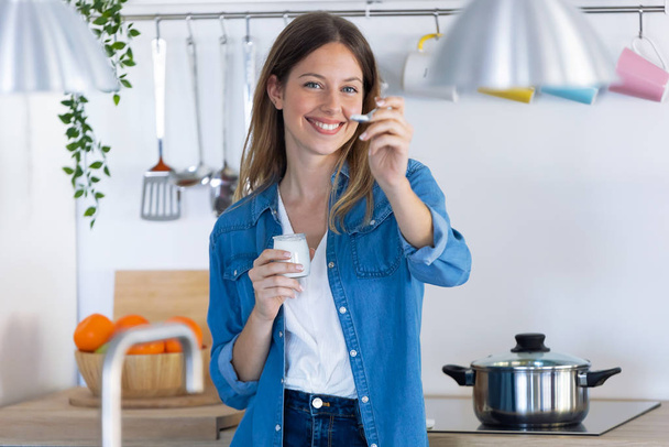 Pretty young woman looking at camera while eating yogurt in the kitchen at home. - Foto, Bild