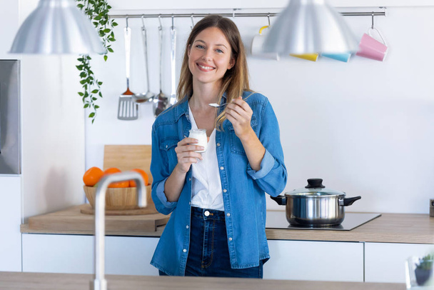 Pretty young woman looking at camera while eating yogurt in the kitchen at home. - Foto, imagen
