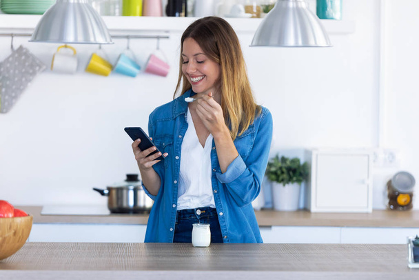 Mulher bonita usando seu telefone celular enquanto come iogurte na cozinha em casa
. - Foto, Imagem