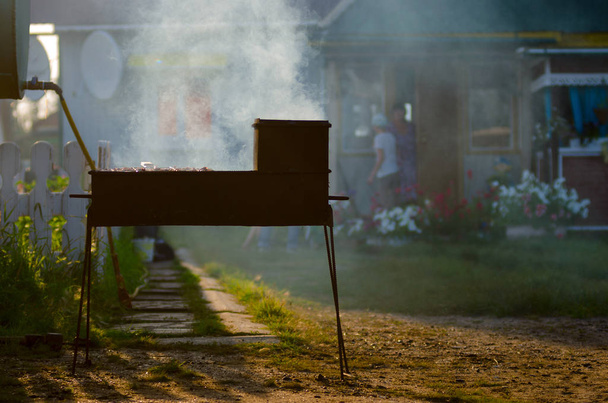 Grade de ferro com churrasqueira fuma nos raios do sol poente no fundo de uma casa privada e mulheres agitadas na distância à porta com flores em Yakutia
. - Foto, Imagem