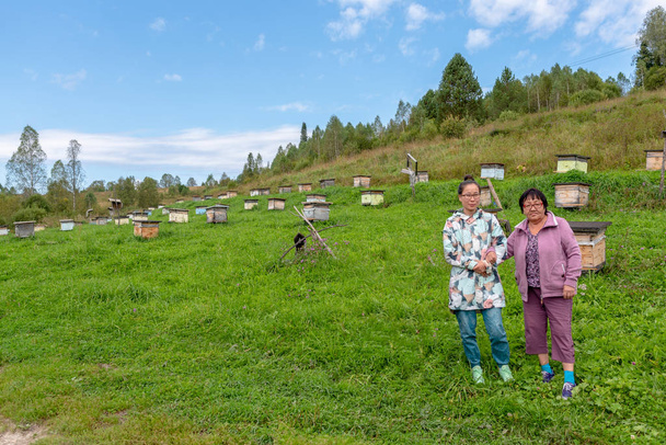 Una familia asiática Yakut una madre anciana y una hija joven posan mano a mano sobre el fondo de colmenas para abejas en un colmenar en las montañas Altai
. - Foto, imagen