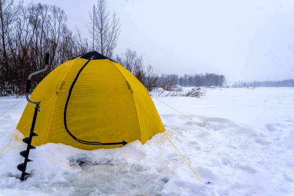 Una tenda da pesca gialla invernale si trova accanto a un impacco di ghiaccio su neve e ghiaccio in una bufera di neve sul lago vicino agli alberi
. - Foto, immagini