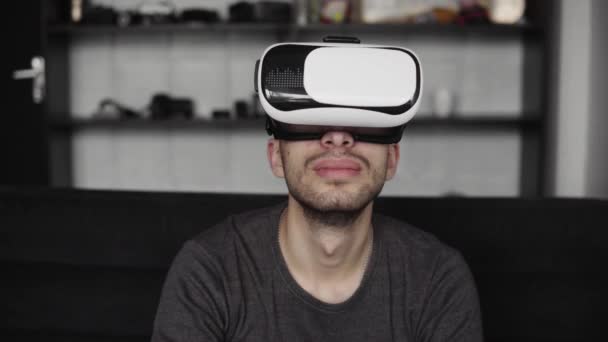 Young bearded man wearing headset of virtual reality in office sitting on a sofa. Exploring VR world and all what he saw in headset. - Video, Çekim