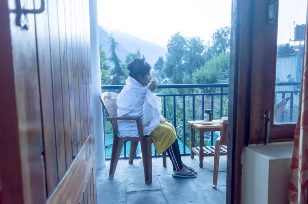 Młoda zamarzająca turystka Podróżująca kobieta owija się w zimową odzież siedząc na wakacyjnym balkonie willi, trzymając rano gorącą filiżankę herbaty. Podróże wakacje aktywne życie i koncepcja zdrowego stylu życia. Mou. - Zdjęcie, obraz
