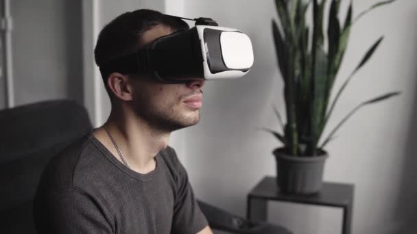 Young bearded man wearing headset of virtual reality in office sitting on a sofa. Exploring VR world and all what he saw in headset. - Metraje, vídeo