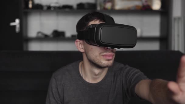 Young bearded man wearing headset of virtual reality in office sitting on a sofa and trying to touch something that he see in virtual world. - Video