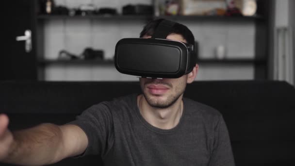 Young bearded man wearing headset of virtual reality in office sitting on a sofa and trying to touch something that he see in virtual world. - Imágenes, Vídeo