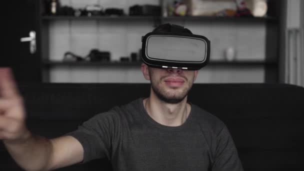 Young bearded man wearing headset of virtual reality in office sitting on a sofa and trying to touch something that he see in virtual world. Using VR headset for work. - Felvétel, videó