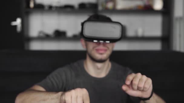Young bearded man wearing headset of virtual reality in office sitting on a sofa and trying to touch something that he see in virtual world. Using VR headset for work. - Séquence, vidéo