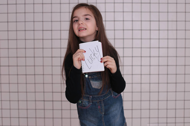 bella ragazza sorridente di otto anni con i capelli lunghi in un dolcevita nero e denim prendisole con un segno "Fortunatamente
" - Foto, immagini