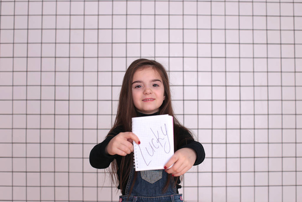 kaunis hymyilevä tyttö kahdeksan vuotta pitkät hiukset musta poolopaita ja denim sundress kanssa merkki "LUCKY
" - Valokuva, kuva