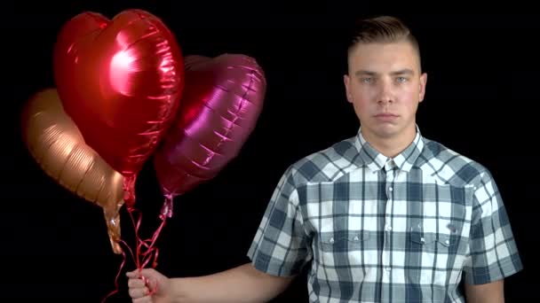 Szomorú fiatalember elengedte a szív alakú lufikat. Egy szomorú ember héliumgolyókkal áll fekete háttérben. Valentin nap a szerelmesek napja.. - Felvétel, videó