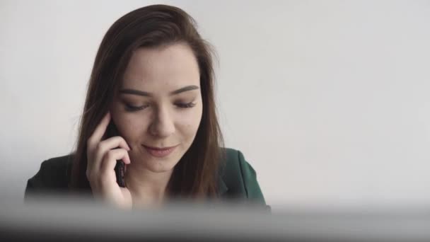 Portrait of young business woman talking phone against computer screen in white office. Female entrepreneur having call and working with computer sitting at table.Telephone work call. - Video, Çekim