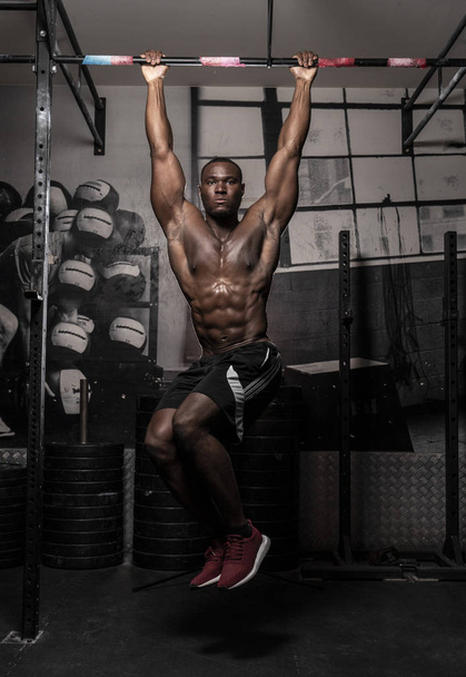 Kaslı, Afrikalı, üstsüz, terli erkek vücut geliştirme sporcusu. Asılı bacak kaldırma hareketi yapıyor. Kaslı, kirli spor salonunda karın kaslarını gösteriyor. - Fotoğraf, Görsel