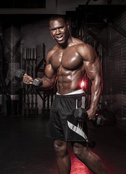 Muscular African American shirtless, sweaty male bodybuilding athlete does barbell curls  in a dark grungy gym with dramatic lighting flare  - Фото, зображення