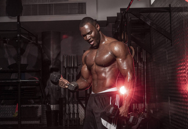 muskulöser afrikanisch-amerikanischer hemdloser, verschwitzter männlicher Bodybuilding-Athlet macht Langhantelcurls in einem dunklen, grungy Fitnessstudio mit dramatischem Lichtkegel  - Foto, Bild