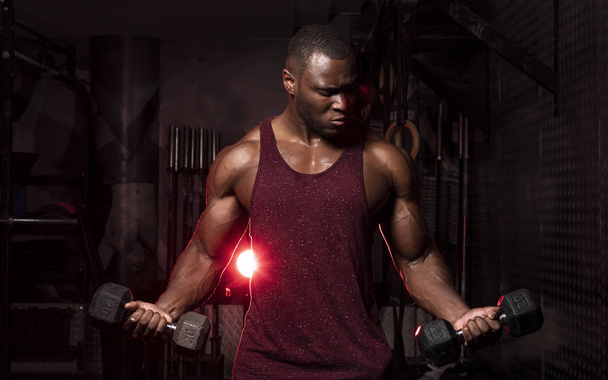 Muskularne Afroamerykanin mężczyzna kulturysta robi hantle loki w ciemnym grungy siłowni z dramatyczne oświetlenie flare  - Zdjęcie, obraz