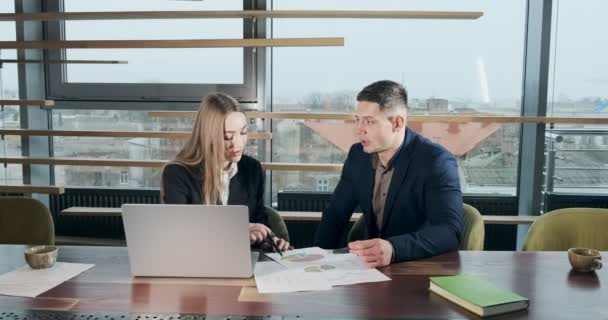 Man and a woman discussing work in the brightly lit modern office. Concerned male and female working with laptop and charts papers at table. Business people discuss project concept. Moving camera - Imágenes, Vídeo