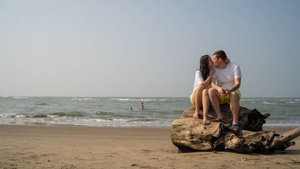 Happy couple sitting on driftwood near sea. Loving couple hugging and touching noses while sitting on driftwood during date on beach against waving sea and cloudless sky - Zdjęcie, obraz