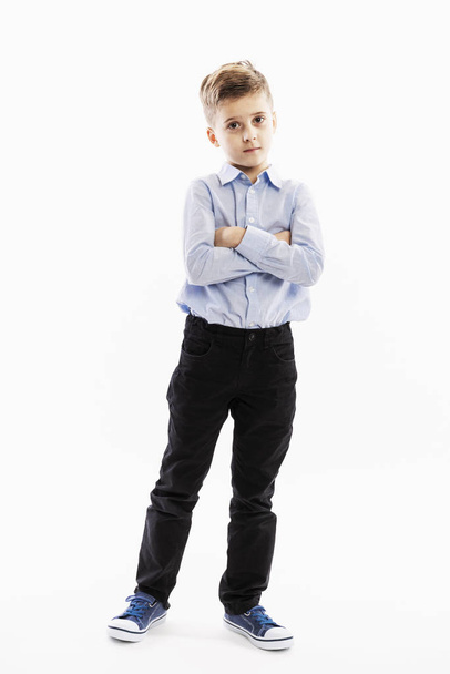 Cute serious boy a schoolboy of 8 years in a blue shirt and trousers stands with his arms crossed on his chest. Full height. White background.  - Фото, изображение