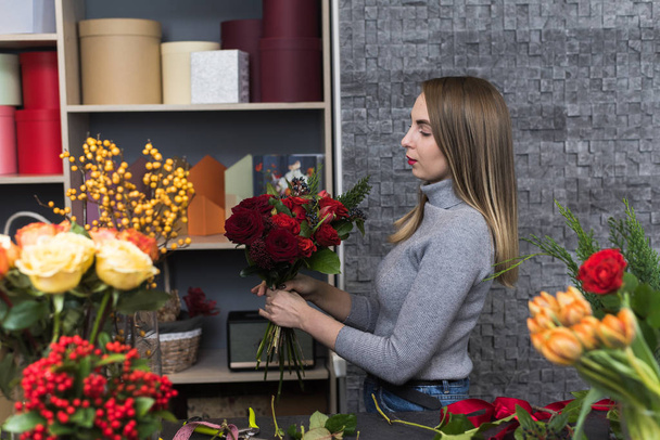 Piękna dziewczyna zbiera ciemny bukiet czerwonych róż w swoim studio. Mężczyzna kwiaciarnia tworząc piękny bukiet w kwiaciarni. Kwiaty dostawy, tworząc zam? wienie, ma? a firma. - Zdjęcie, obraz