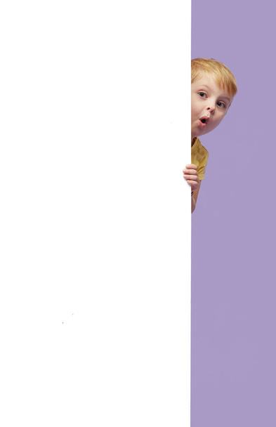 Little smiling child standing behind a white blank panel isolated against purple background. Peeking out from behind a banner, empty space for text. - Photo, Image