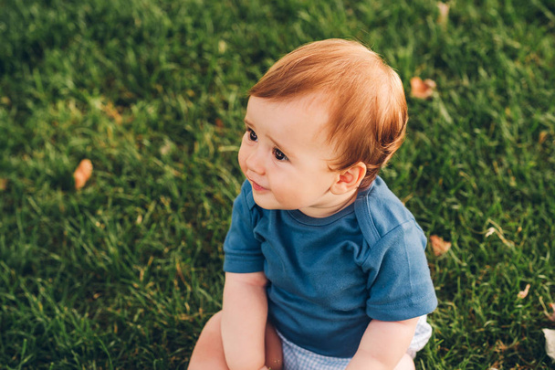 Portrait extérieur d'adorable petit garçon roux jouant dans le parc d'été, assis sur l'herbe verte, portant un body bleu
 - Photo, image