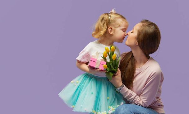 8 march card! Little daughter kisses and hugging her mother with yellow flowers tulip and gift box. Purple studio background. Mothers day concept - Фото, изображение