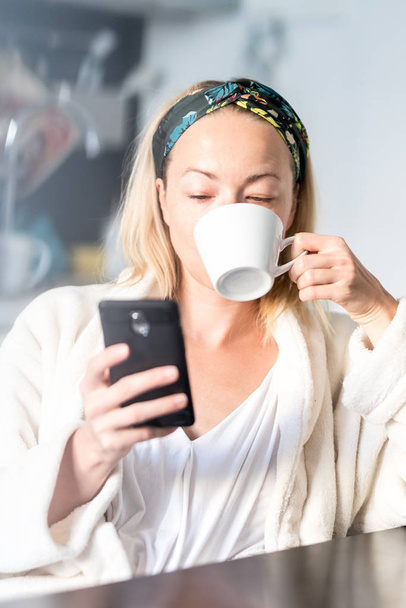 Schöne kaukasische Frau zu Hause, fühlt sich wohl im weißen Bademantel, nimmt sich Zeit für sich, trinkt Morgenkaffee und liest morgens Nachrichten auf dem Handy - Foto, Bild